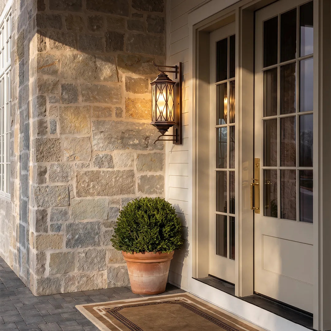 Bronze outdoor sconce mounted on a stone exterior wall beside a glass door, blending classic style with warm illumination.