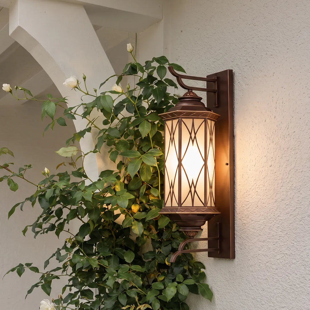 Bronze outdoor sconce fixed to a white exterior wall, showcasing a traditional lantern design beside climbing greenery.