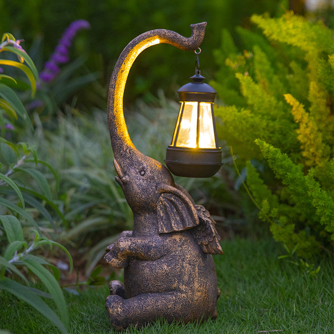 Brown resin elephant garden light fixture with illuminated lantern positioned in lush green landscaping