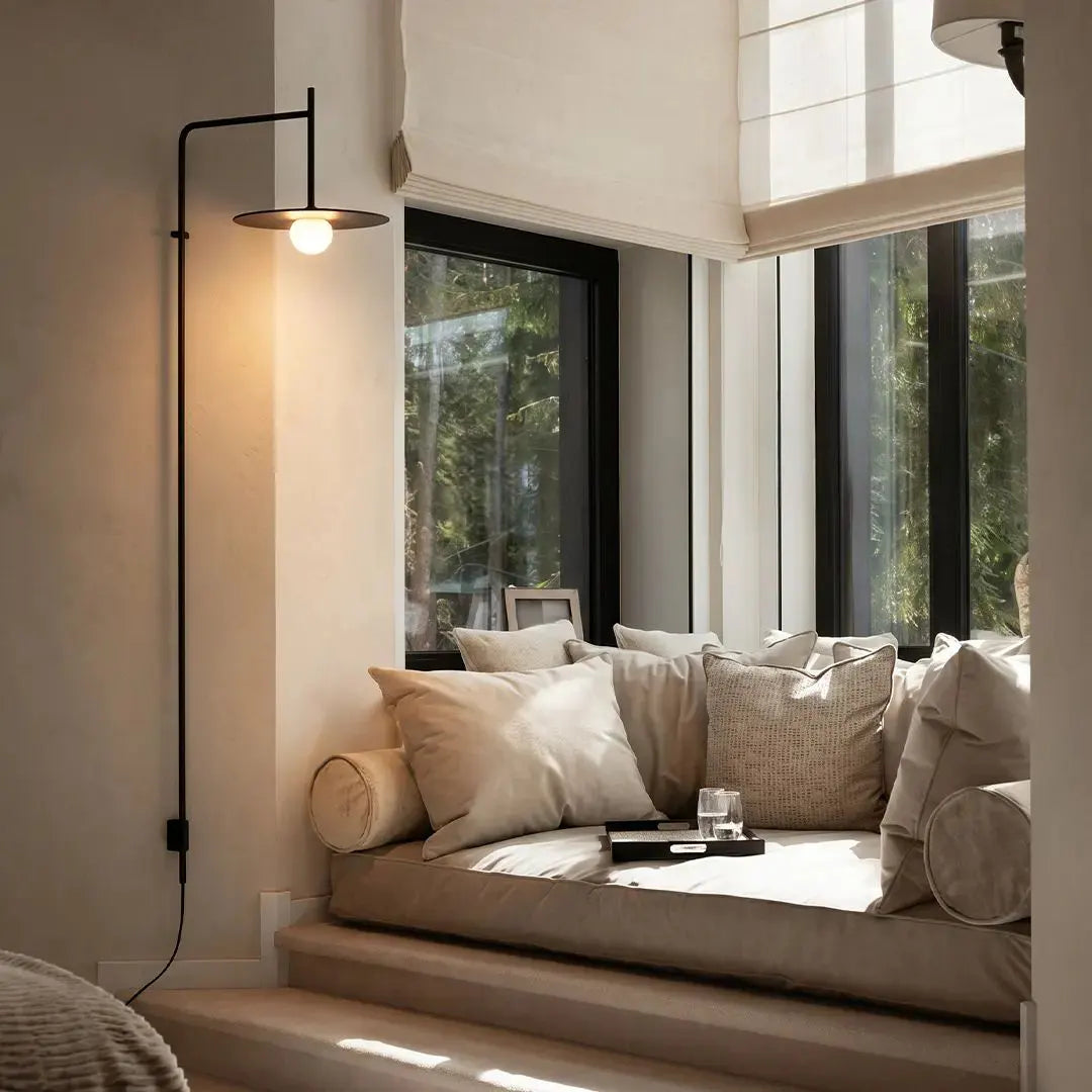 Cantilever wall lamp mounted beside a window seat, casting warm light in a cozy neutral room.