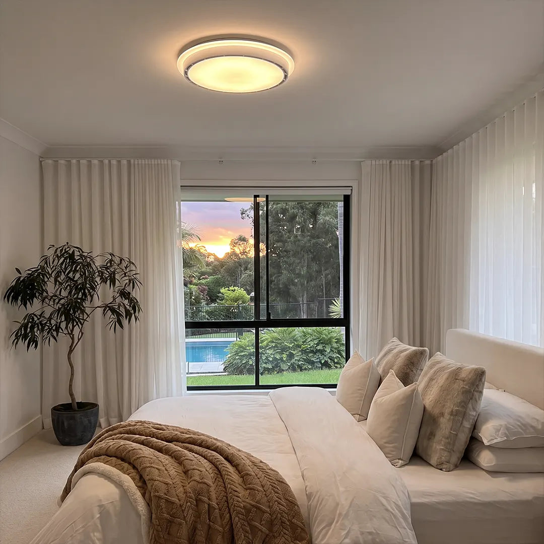 Ceiling light with hidden fan mounted in a bright bedroom with large windows, white curtains, and a neutral bed.