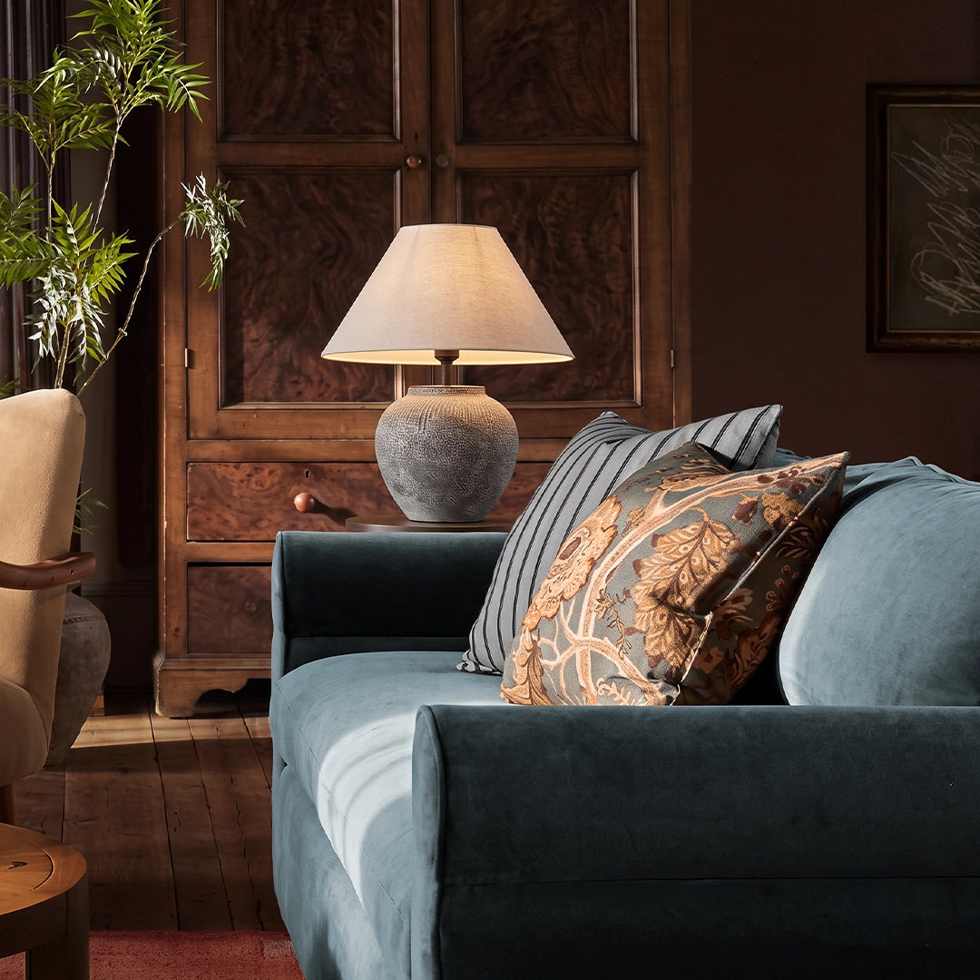 Ceramic base table lamp beside a blue sofa, featuring a textured gray ceramic base and tapered fabric shade.