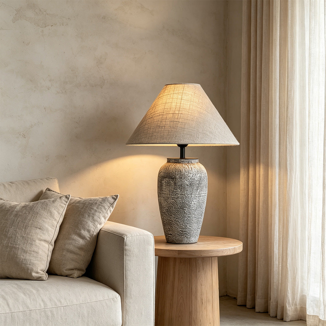 Ceramic base table lamp on a round wood side table next to a neutral sofa in a soft-toned living room.