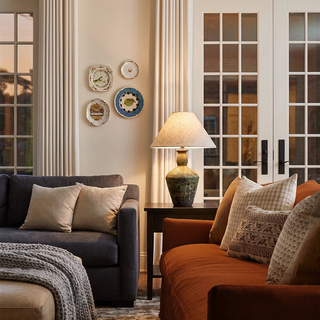 Ceramic bedside table lamp styled between two sofas in a cozy living room, with a patterned ceramic base and beige fabric shade.