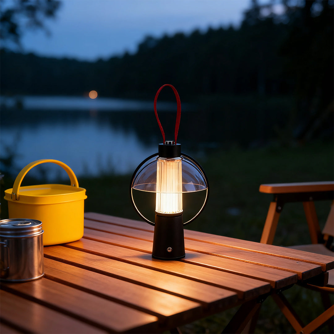 The warm glow of a circle table lamp with clear glass shade placed on a wooden table near a tranquil lake at dusk. 