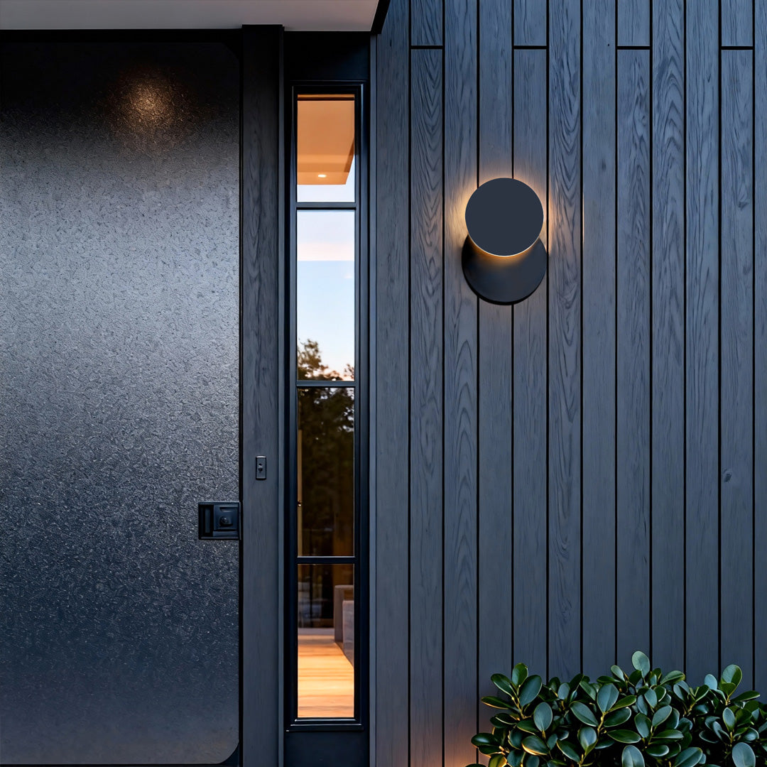 Modern circular outdoor lighting sconce installed on a dark vertical panel exterior, creating soft warm ambient light.