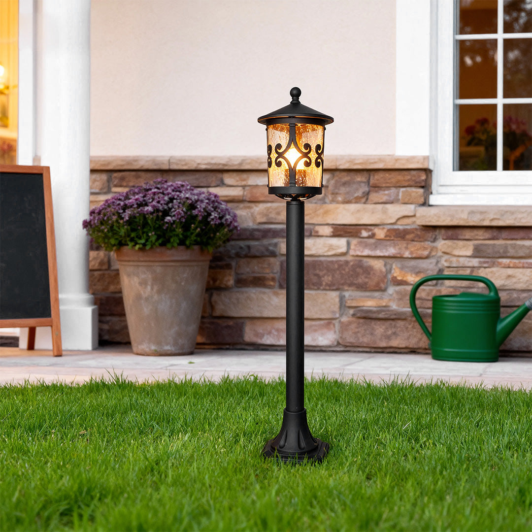 Classic bollard LED light positioned on front yard grass near brick patio with potted purple flowers