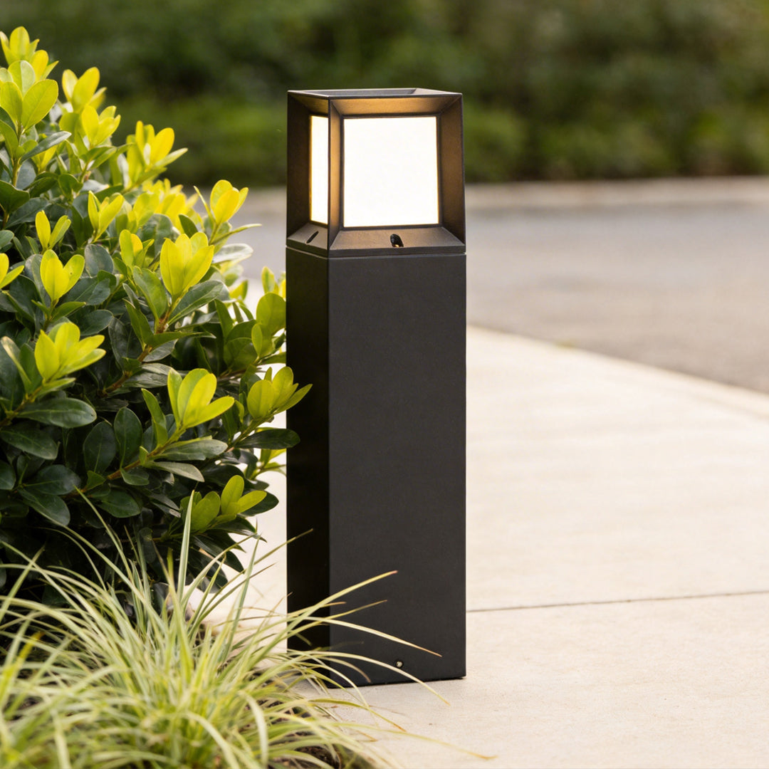 Close up detail of a black outdoor bollard light installed next to green bushes on a sunny day.