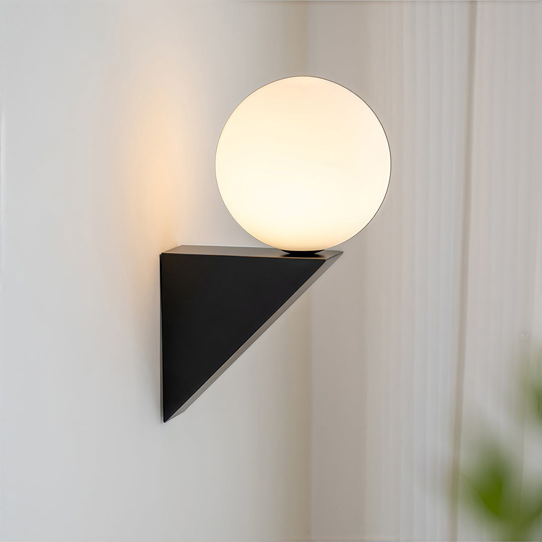 Close-up of a modern globe wall light featuring a frosted white sphere resting on a black triangular base, illuminating a white wall.