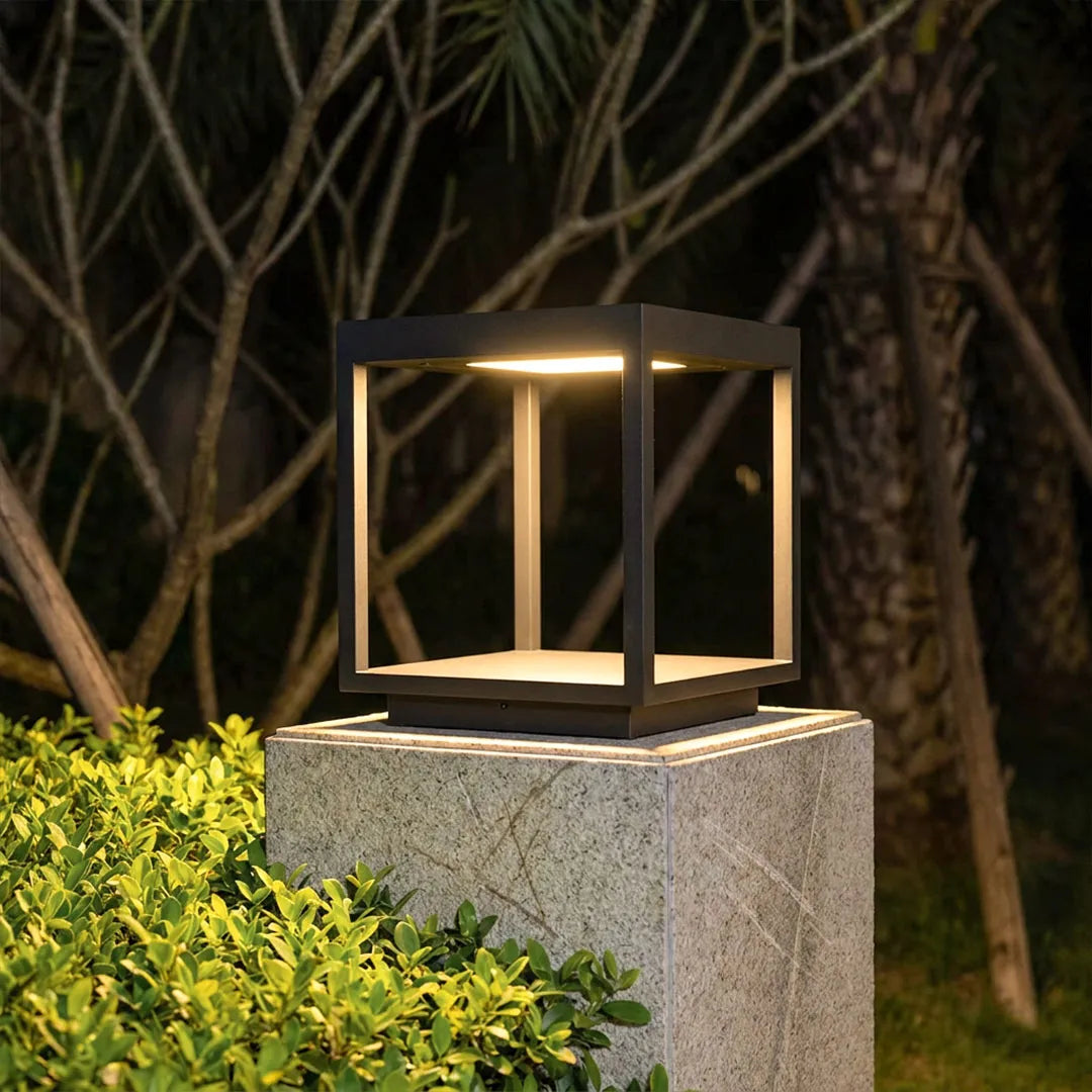 Concrete pillar light glowing on a square post surrounded by garden greenery at night.