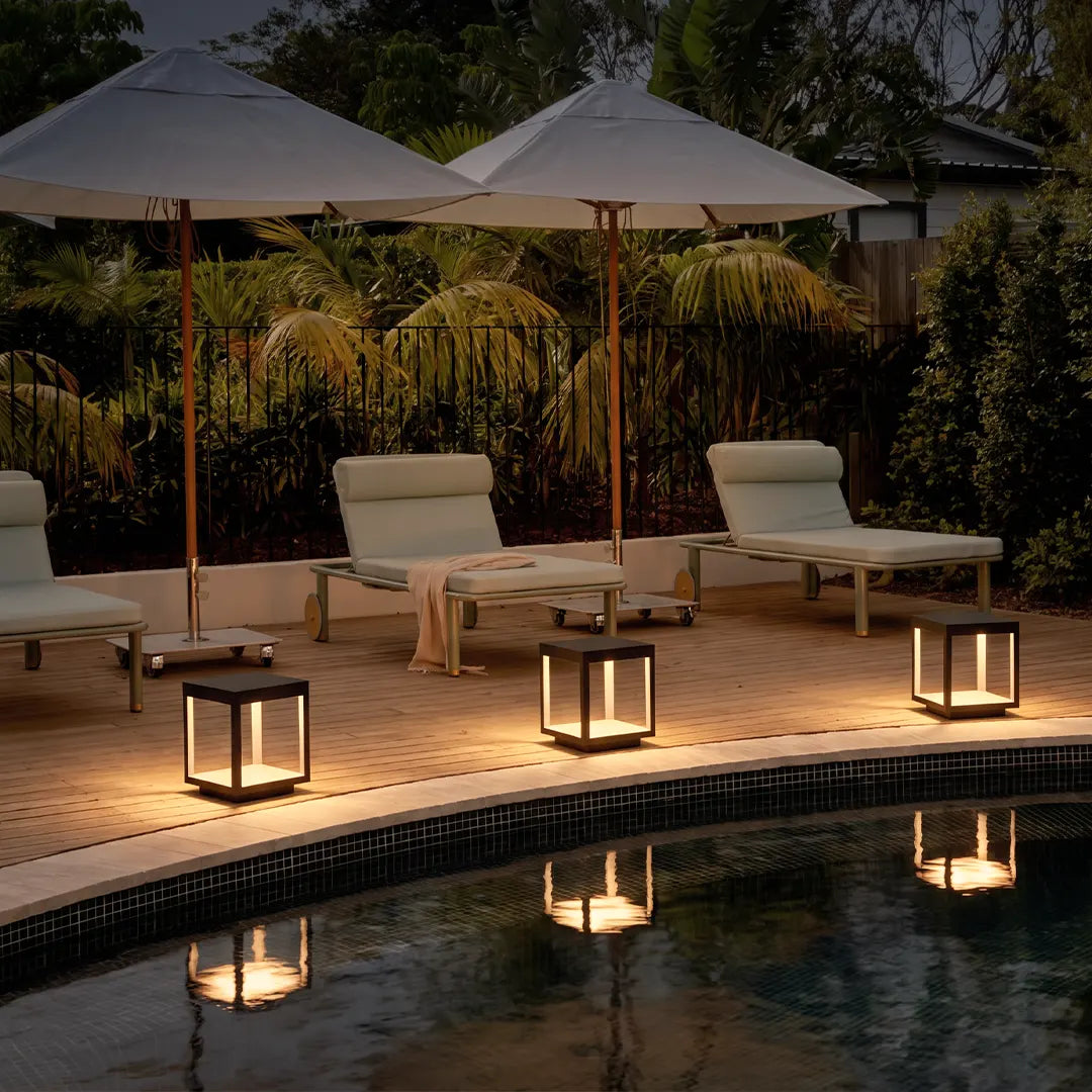 Concrete pillar lights illuminating a poolside patio with lounge chairs and umbrellas in a modern outdoor setting.