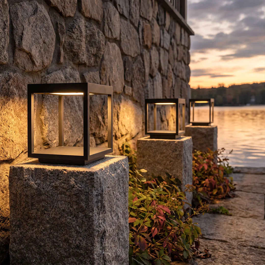 Concrete pillar lights installed on stone columns beside a waterfront garden path at dusk.
