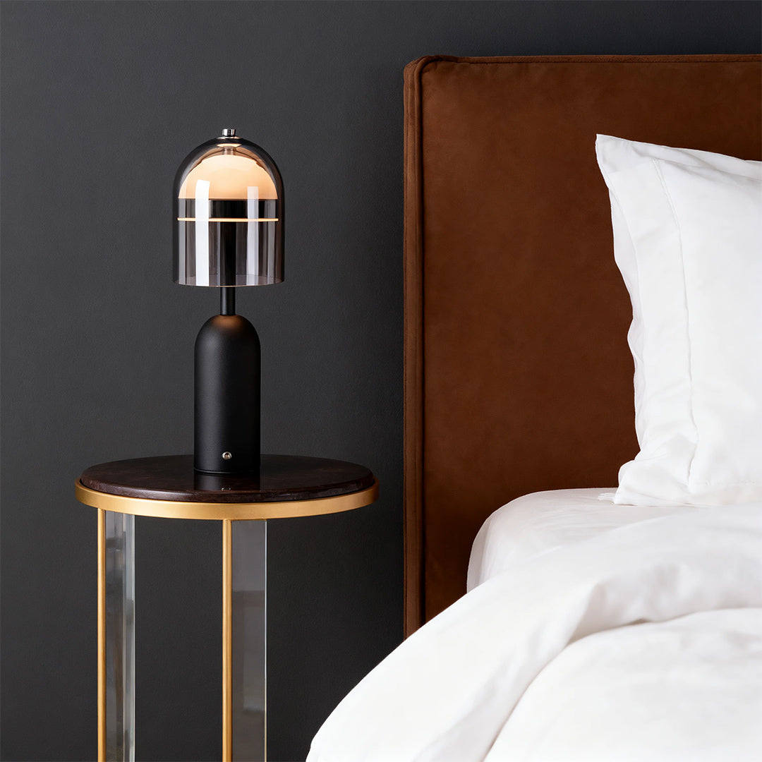Contemporary black glass shade table lamp on a gold and glass side table beside a bed with a brown leather headboard. 