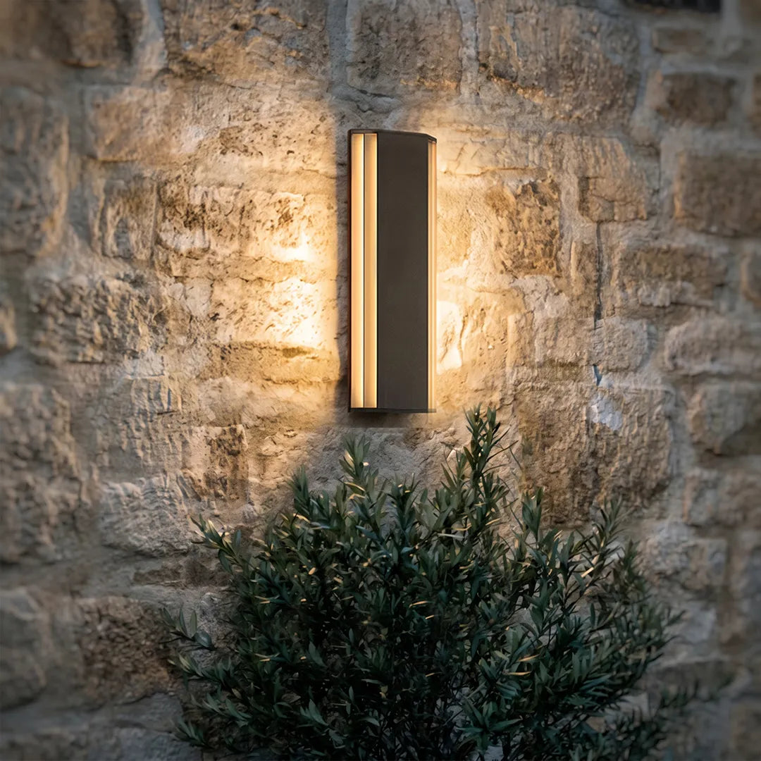 Contemporary LED wall light mounted on a rustic stone wall above landscaping, providing warm outdoor accent lighting.