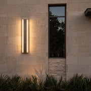 Contemporary LED wall light mounted on a beige stone exterior wall beside a narrow window, casting a warm ambient glow.