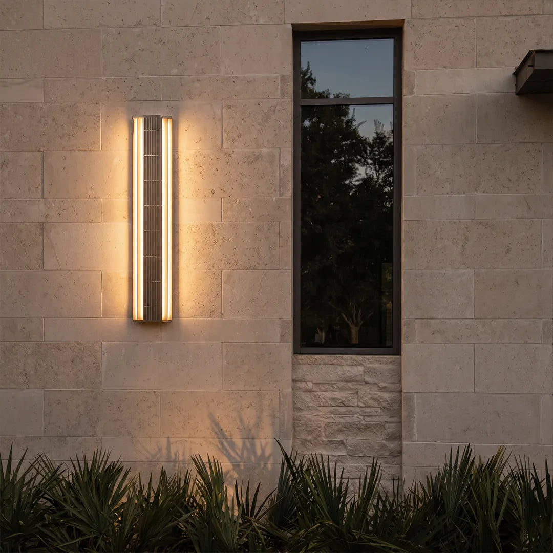 Contemporary LED wall light mounted on a beige stone exterior wall beside a narrow window, casting a warm ambient glow.