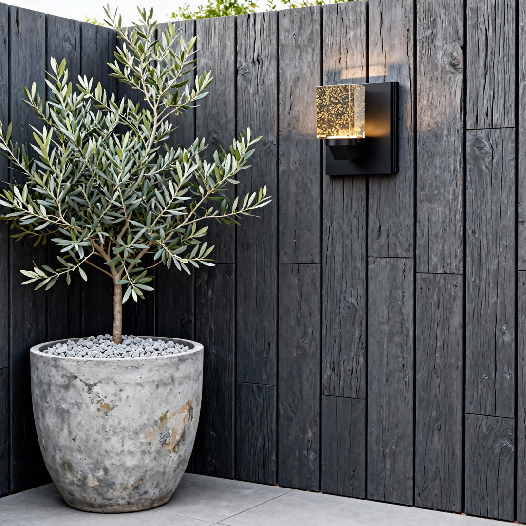 Contemporary outdoor wall lighting crystal sconce mounted on weathered wood fence beside potted olive tree