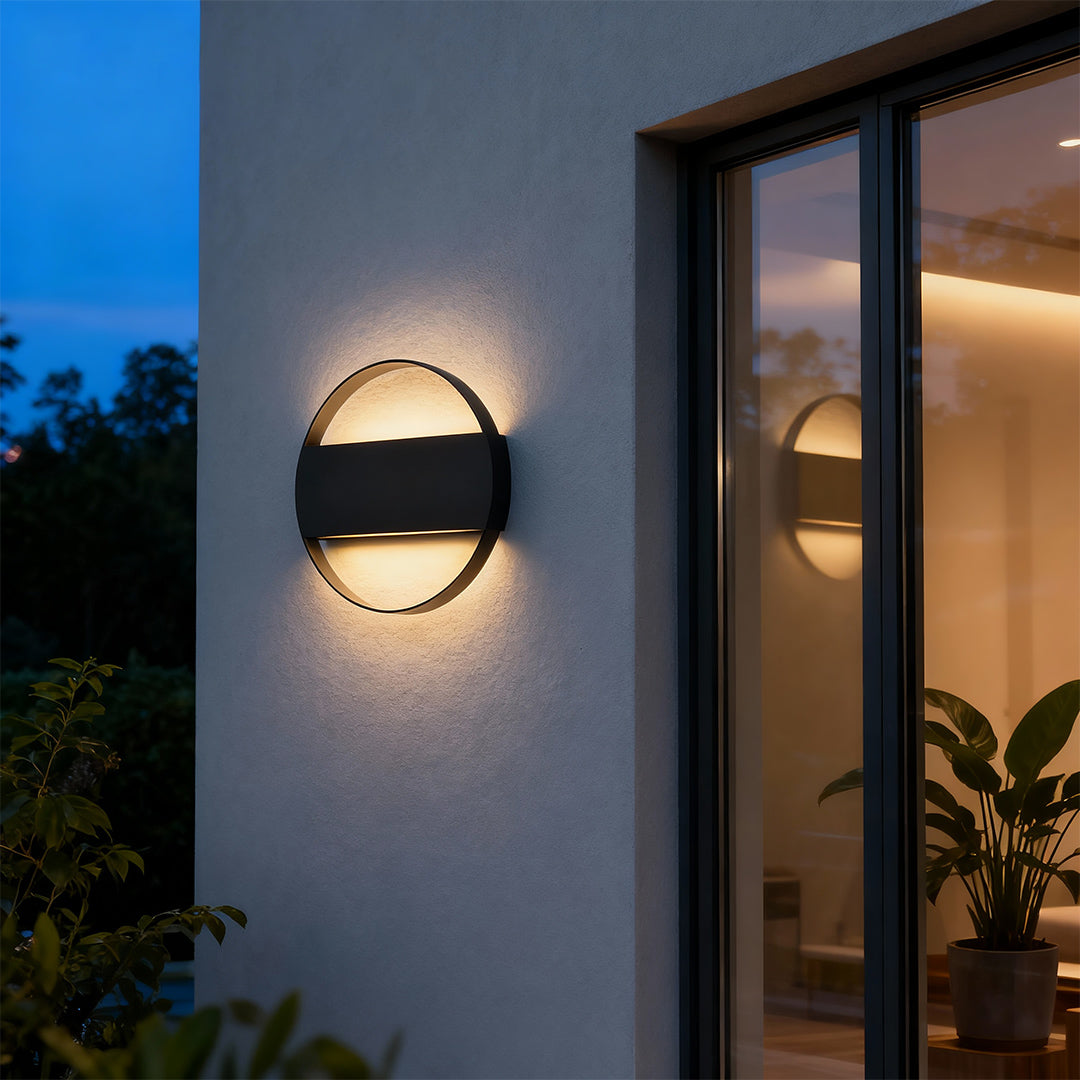 Contemporary round wall mount exterior light with glass shade chandelier aesthetic illuminating modern gray exterior wall at dusk.