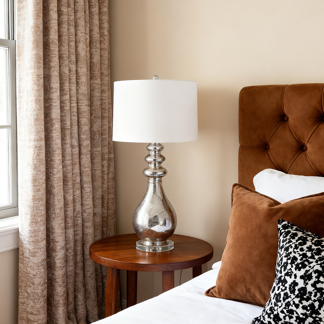 Cozy bedroom scene with a stylish modern silver table lamp resting on a round wood nightstand beside a tufted brown headboard.