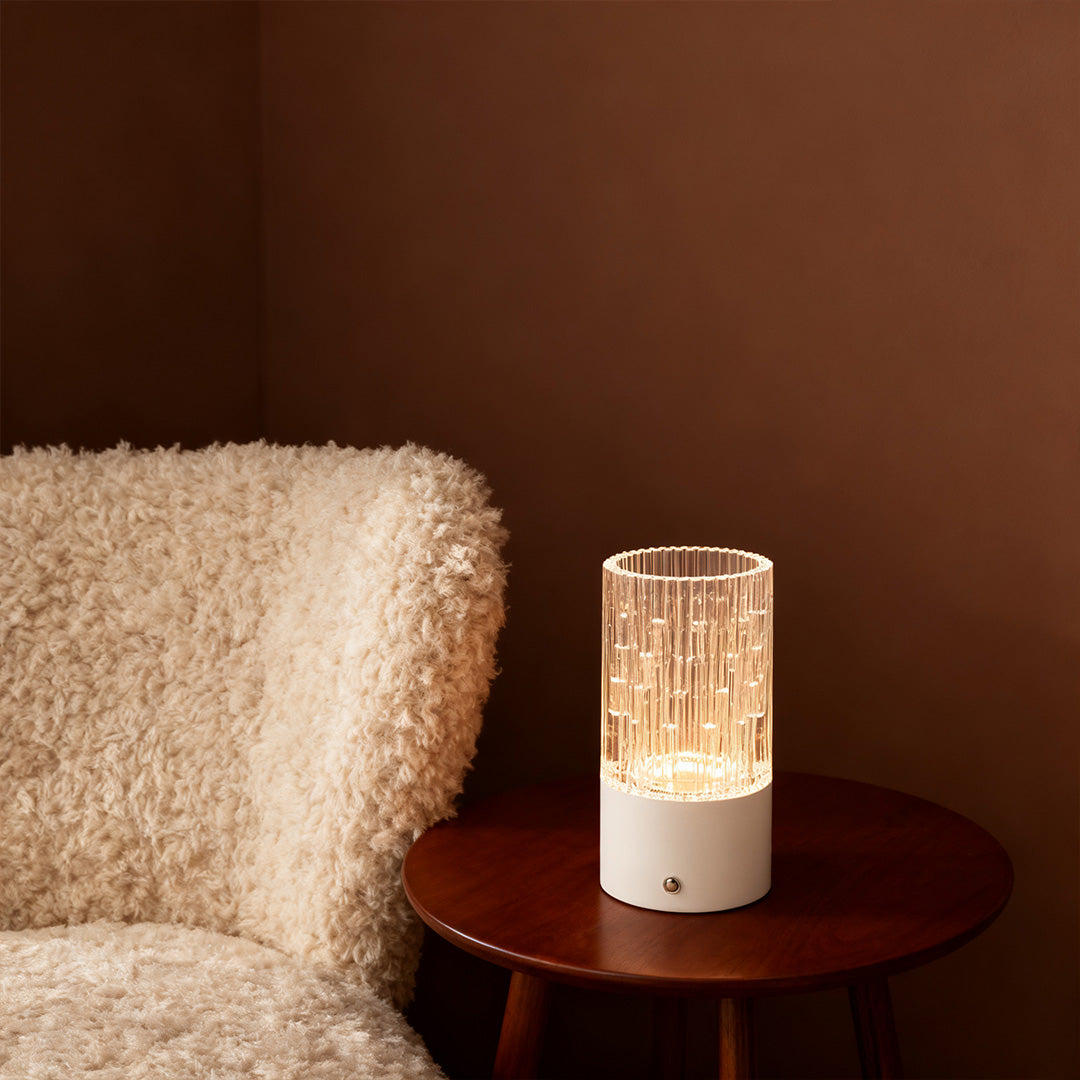 Cozy white deco table lamp with crystal effect cylinder resting on a small round wooden table next to a plush armchair.