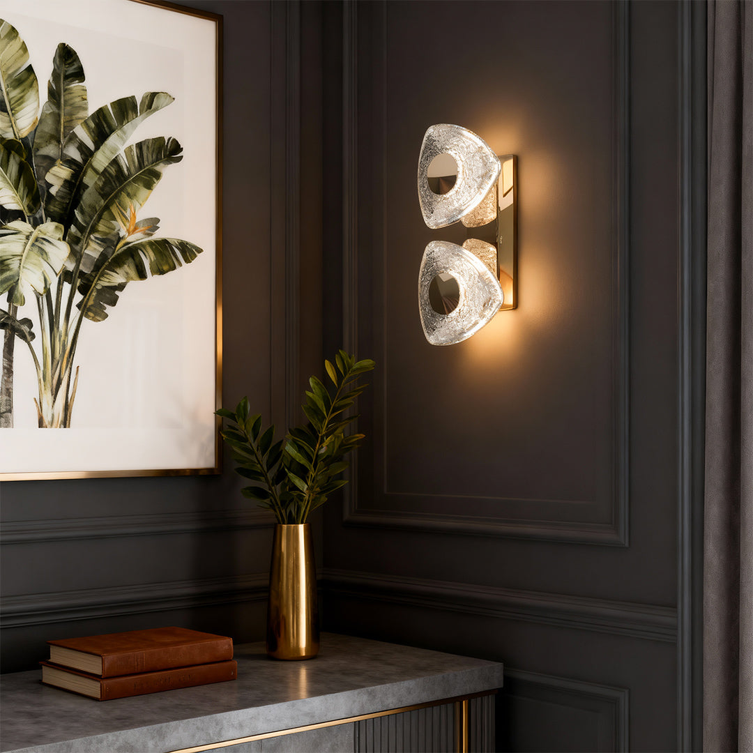 Crystal wall sconce light fixture mounted on dark paneled wall beside modern console table with botanical accents