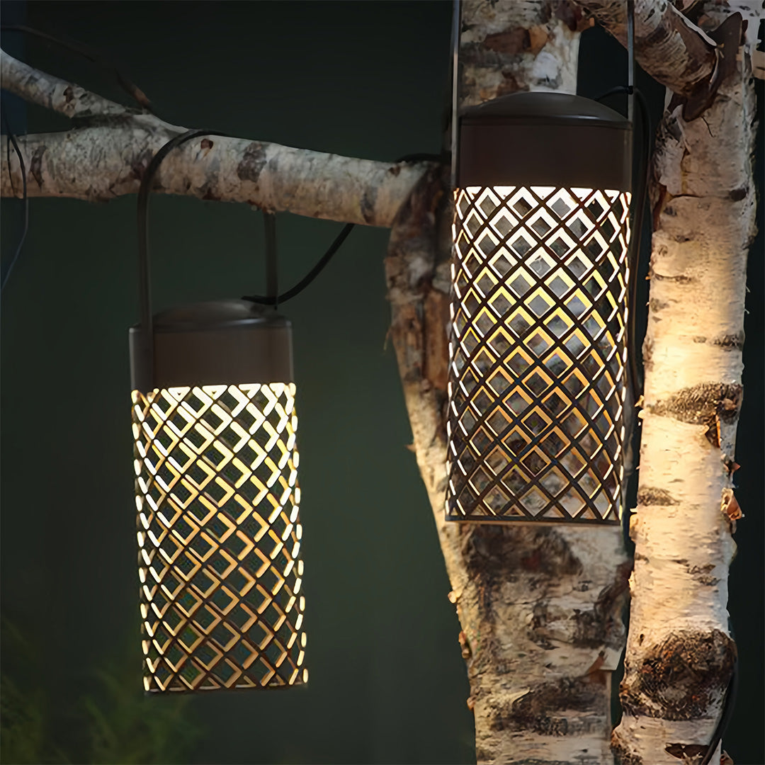Cylindrical black outdoor pendant lantern with diamond cutout screen, hung from a birch tree trunk, casting a decorative shadow.