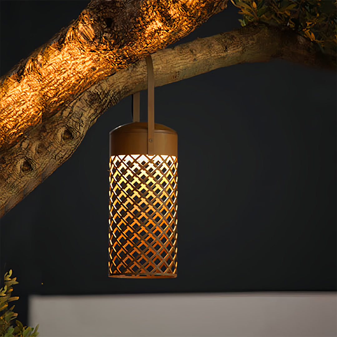 Cylindrical black outdoor pendant lantern with diamond lattice pattern, hanging from a tree branch in warm golden light.