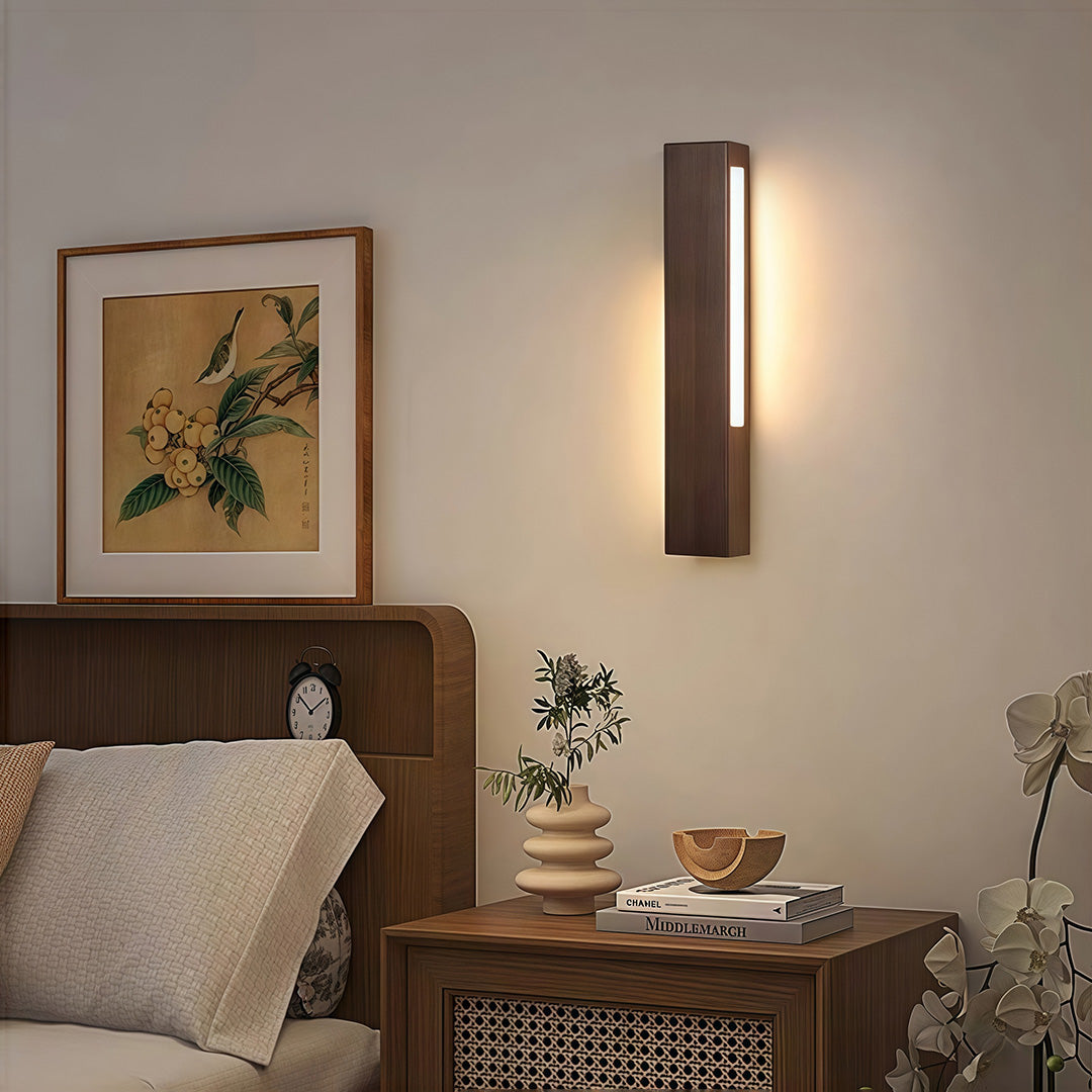 Dark wood vertical LED Japanese wall light providing soft ambient illumination above a traditional bedside table in a neutral room.