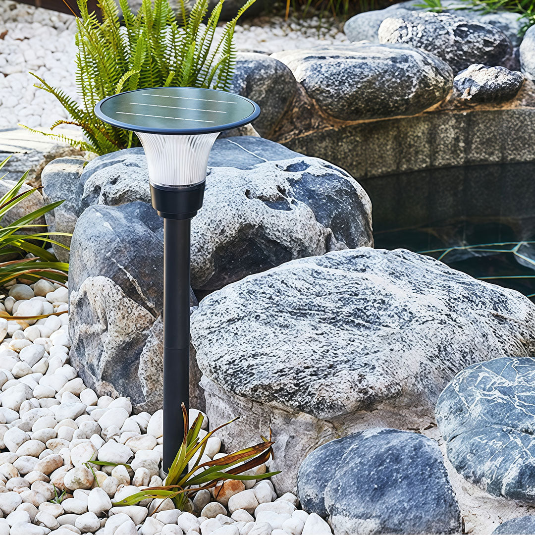 Daytime view of the black solar pathway lights stake installed in a gravel-and-rock garden by a pond.