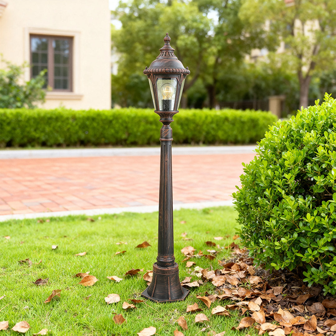 Decorative bollard light standing on lawn edge beside brick pavers with green hedges and building