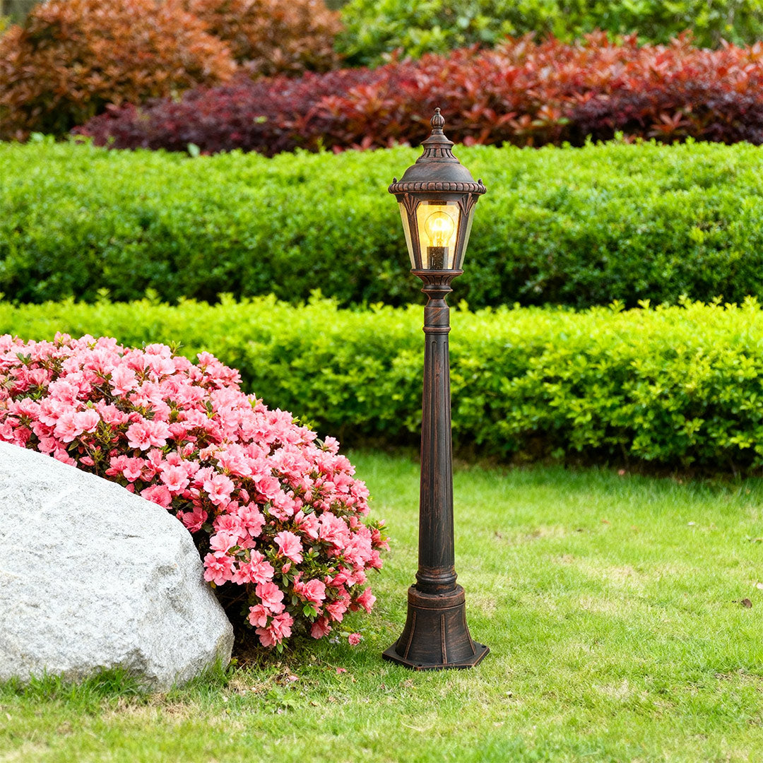 Decorative bollard light standing on white stone border near pink flowers with layered green hedges