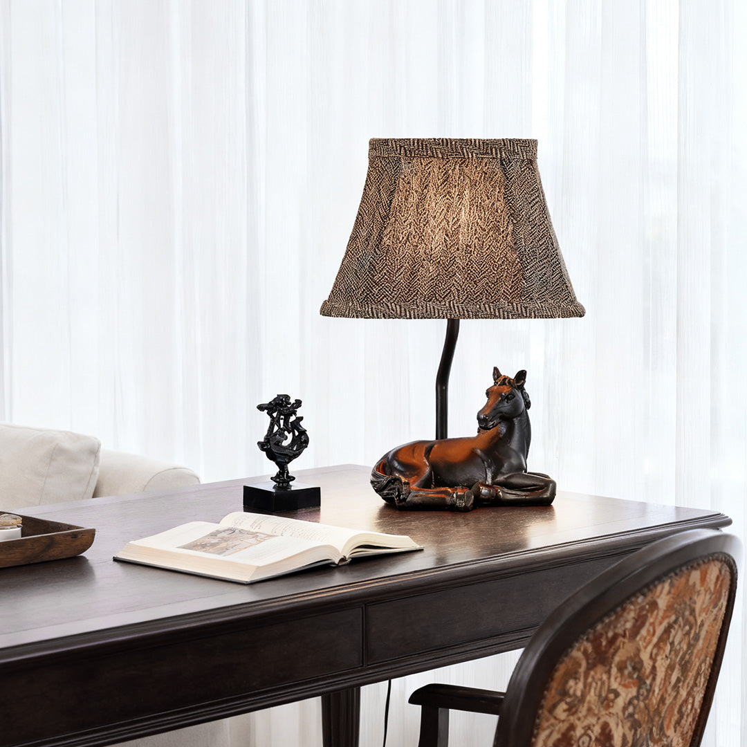 Decorative table lamp horse used as a focal point on a vintage wooden writing table in front of sheer white curtains.