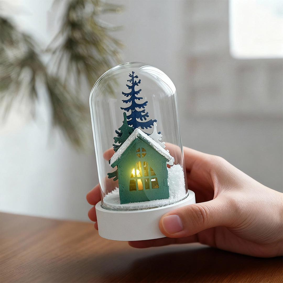 Hand holding small decorative led table lamp with glass cover containing miniature lit cottage and winter landscape