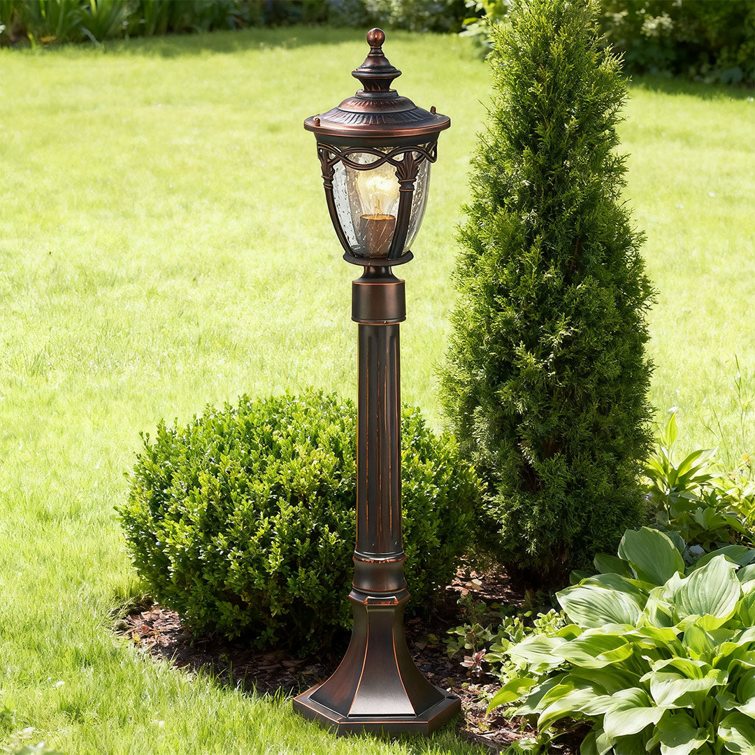 Decorative outdoor light bollard standing among manicured round bushes on bright green grass