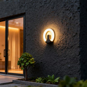 Designer black wall sconce light with glass shade chandelier aesthetic creating beautiful ring of light on dark textured exterior.