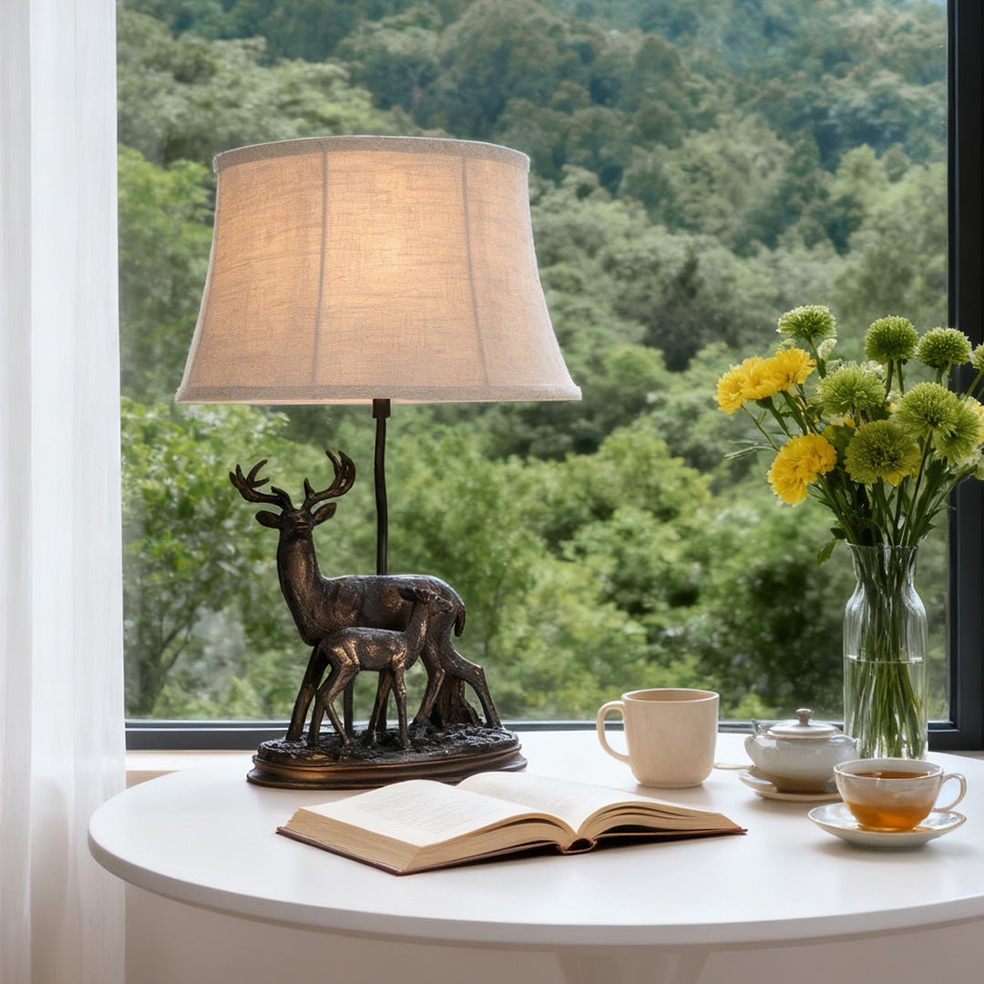 Decorative deer table lamp placed on a white table with a scenic forest view, highlighting the detailed stag and fawn sculpture.