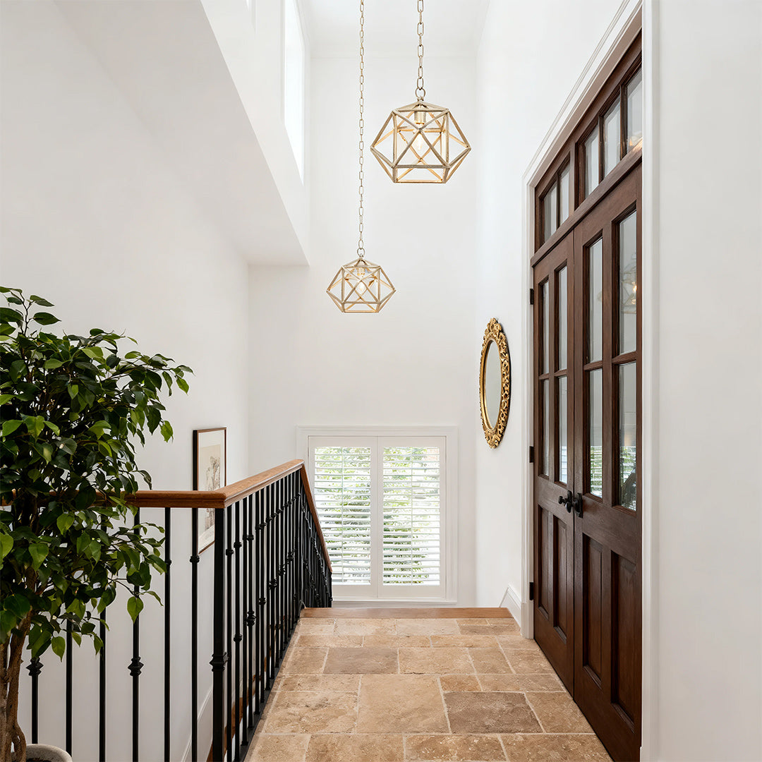 Gold metal pendant light hanging in a double-height entryway, casting light onto a classic wrought iron staircase railing and stone floor.