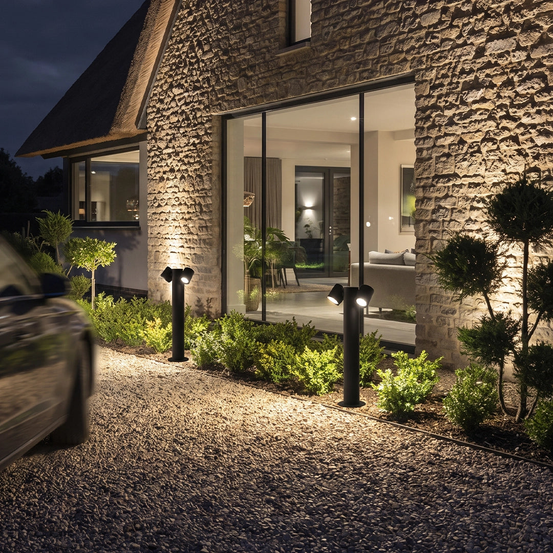 Double light bollard lights lining a gravel driveway beside a modern stone house at night.