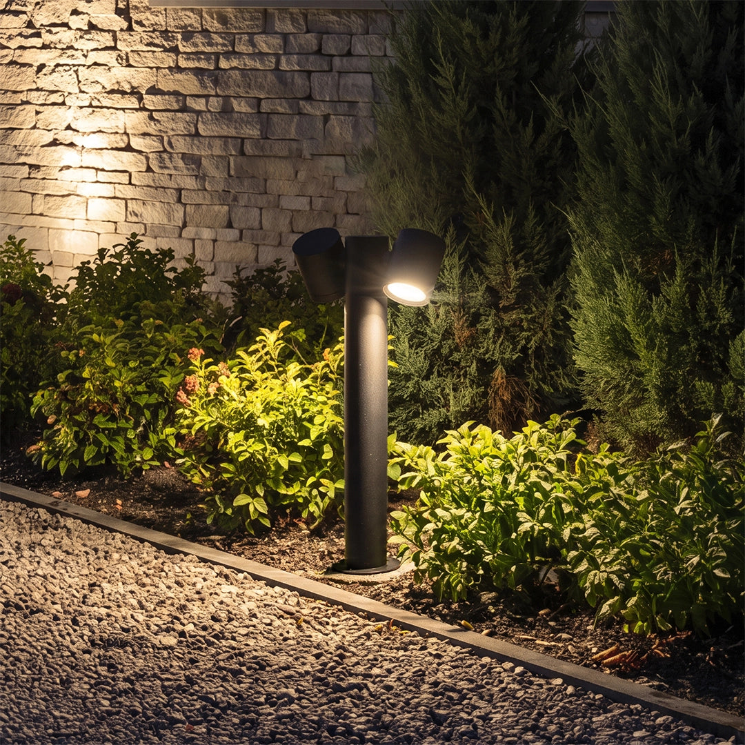Double light bollard light installed beside a garden bed, casting warm light on plants and stone walls.