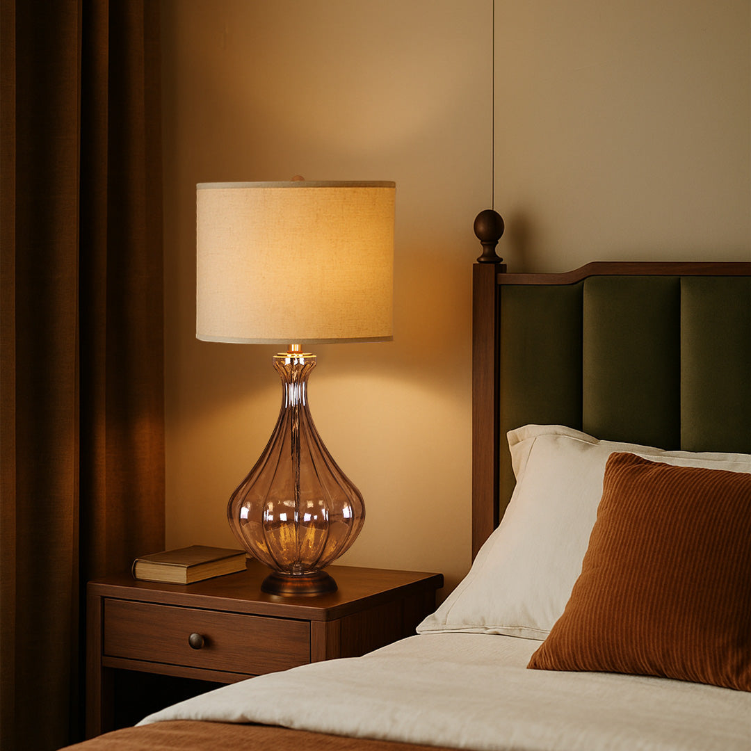 Elegant glass base table lamp with an antique copper finish and white fabric drum shade, placed on a dark wood nightstand.