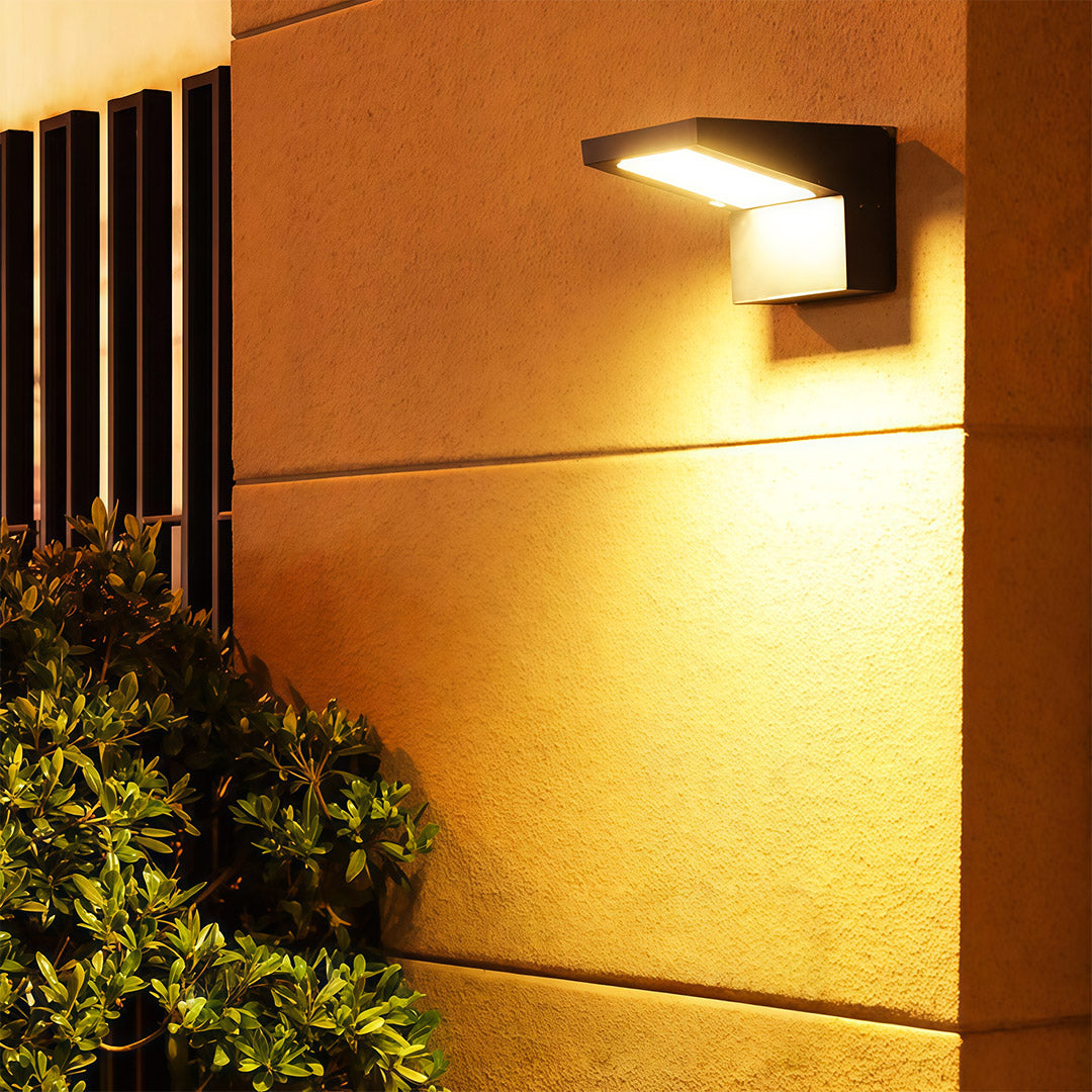 Elegant black wall mount outdoor light fixture providing warm accent lighting above a bush on a textured wall.
