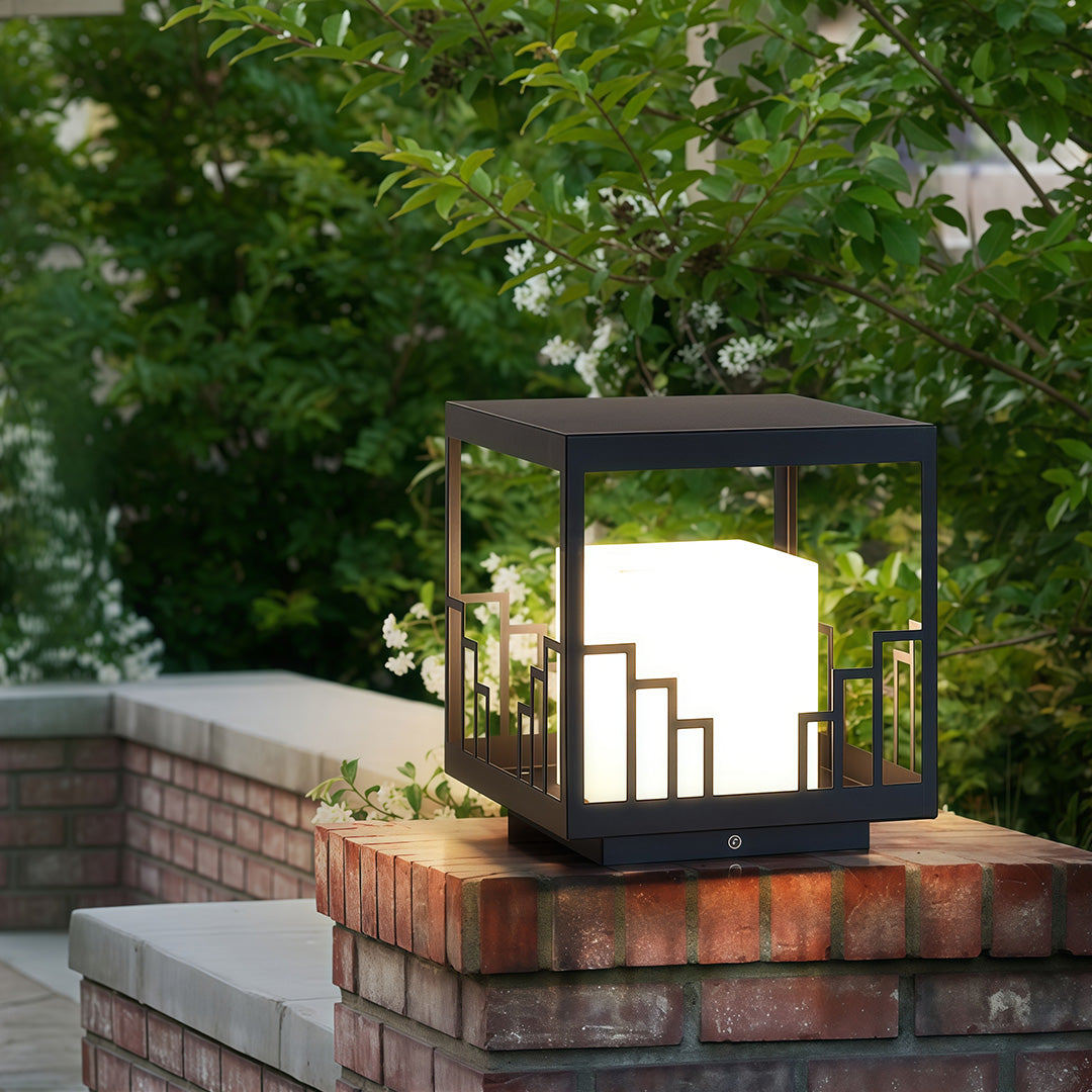 Elegant outdoor pillar light featuring glass shade chandelier styling illuminating brick entrance pillar in garden setting at dusk.