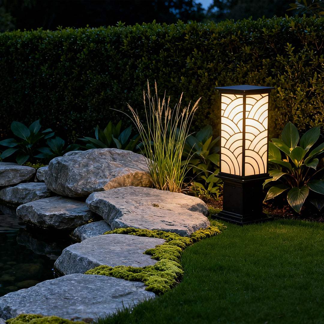 Exterior bollard light placed among stones and garden plants, casting warm illumination across a landscaped walkway.