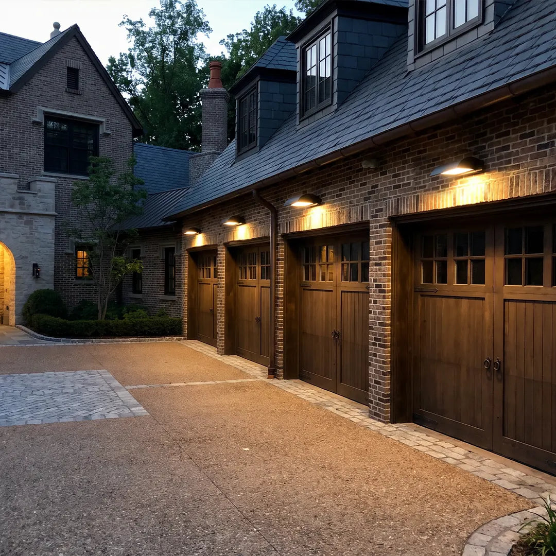 Exterior down wall lights mounted above dark wood garage doors on a large stone house exterior at dusk.