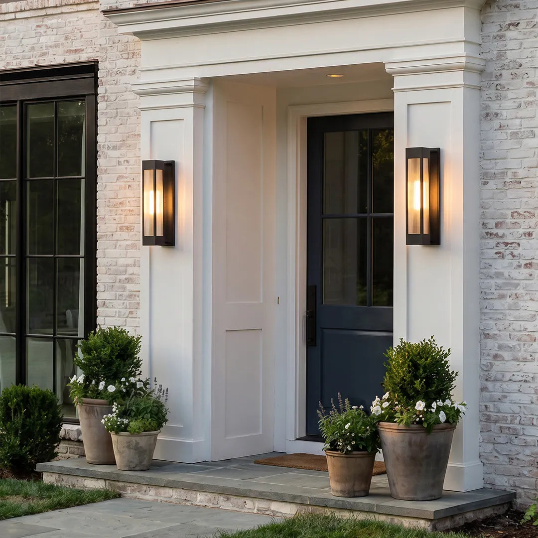 Exterior LED wall sconce lighting flanking a front door on a white brick home with potted plants.