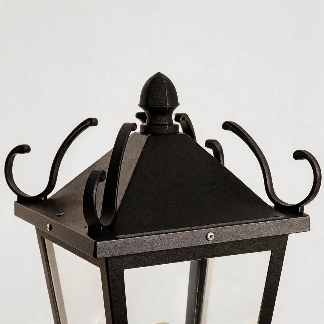 Exterior pillar light detail showing the black lantern top with decorative scroll accents and a textured outdoor finish.
