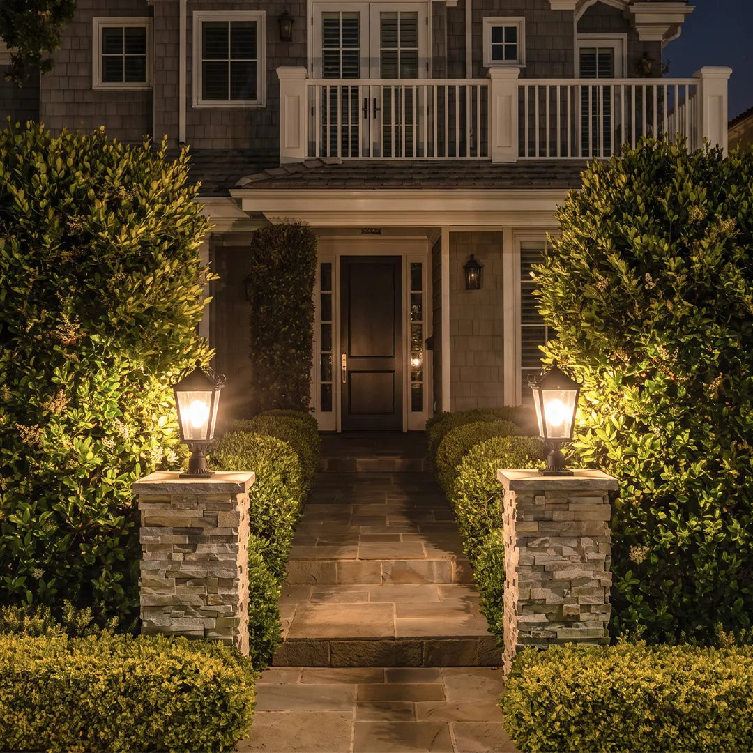 Exterior pillar lights illuminating a front walkway between stone columns and trimmed hedges, creating a warm and inviting entrance.