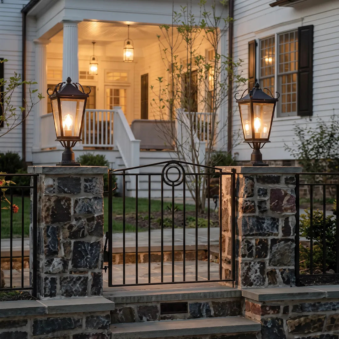 Exterior pillar lights mounted on stone gate posts in front of a white house, enhancing curb appeal with classic outdoor lighting.