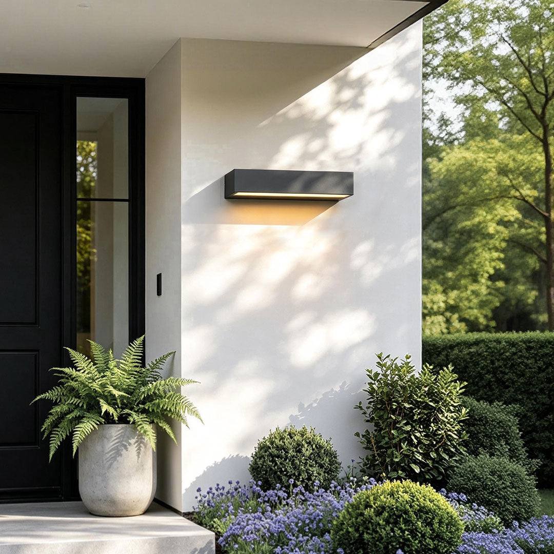 Exterior porch light fixture illuminating a front entry beside a black door and landscaped garden.