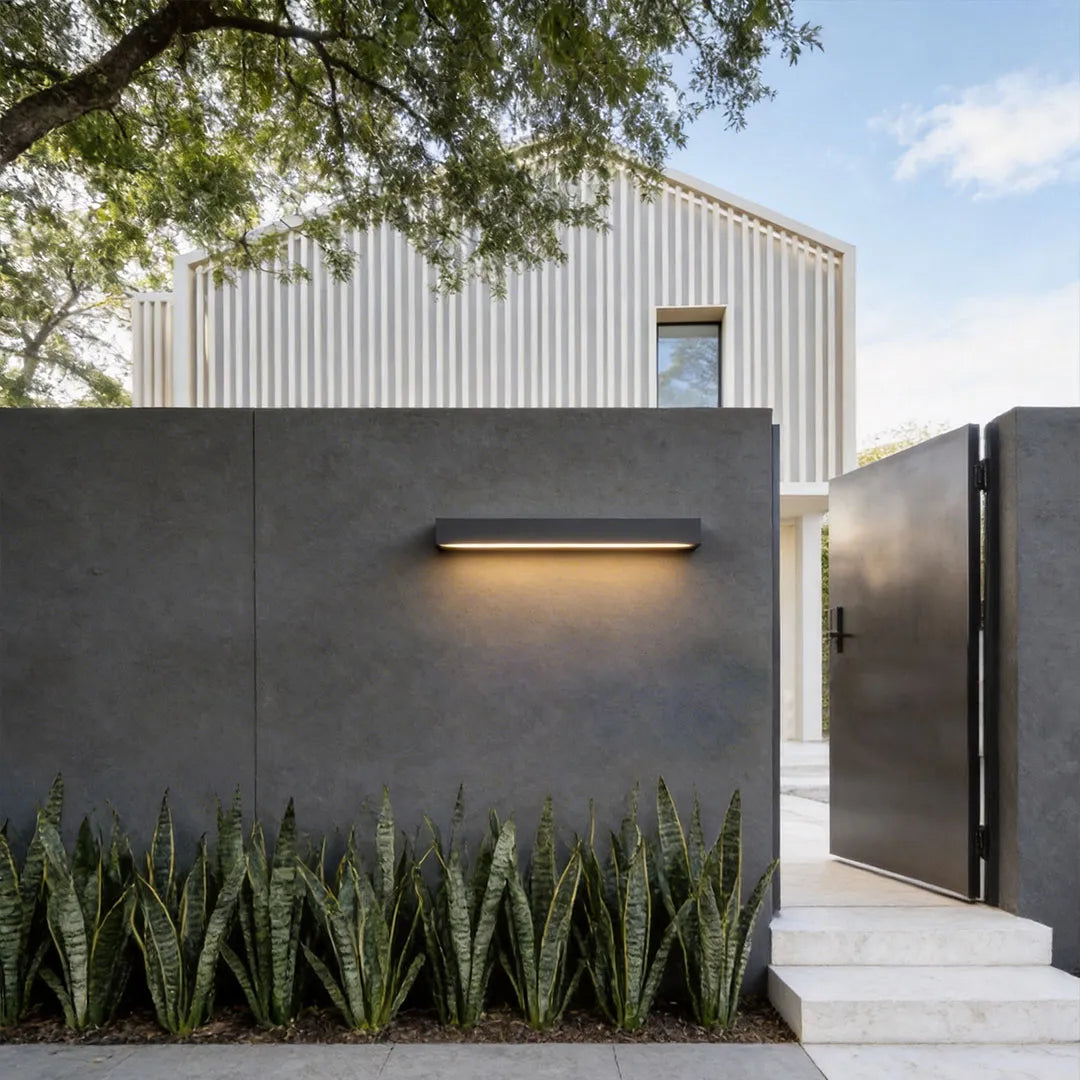 Exterior porch light fixture mounted on a dark modern wall above a row of landscape plants.